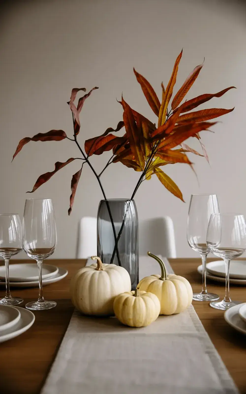 Formal dining table with vases of fall leaves, white pumpkins, place settings, and wine glasses.