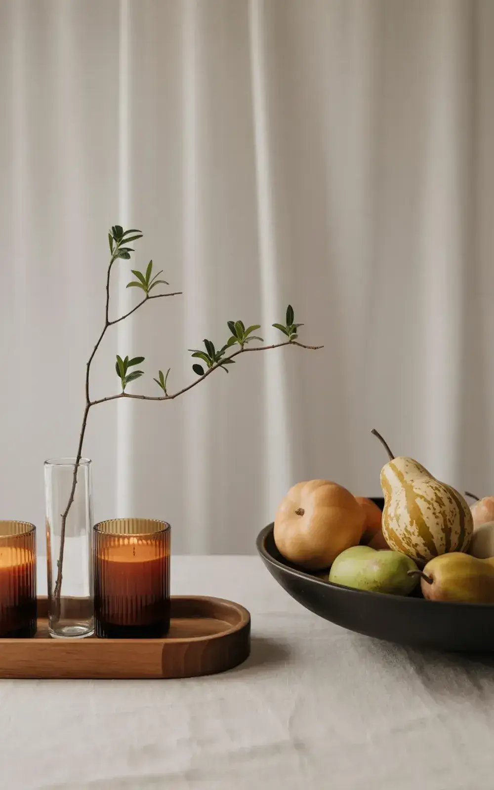 Two lit candles, a branch in a clear vase, and a bowl of mixed fruit on wooden trays.