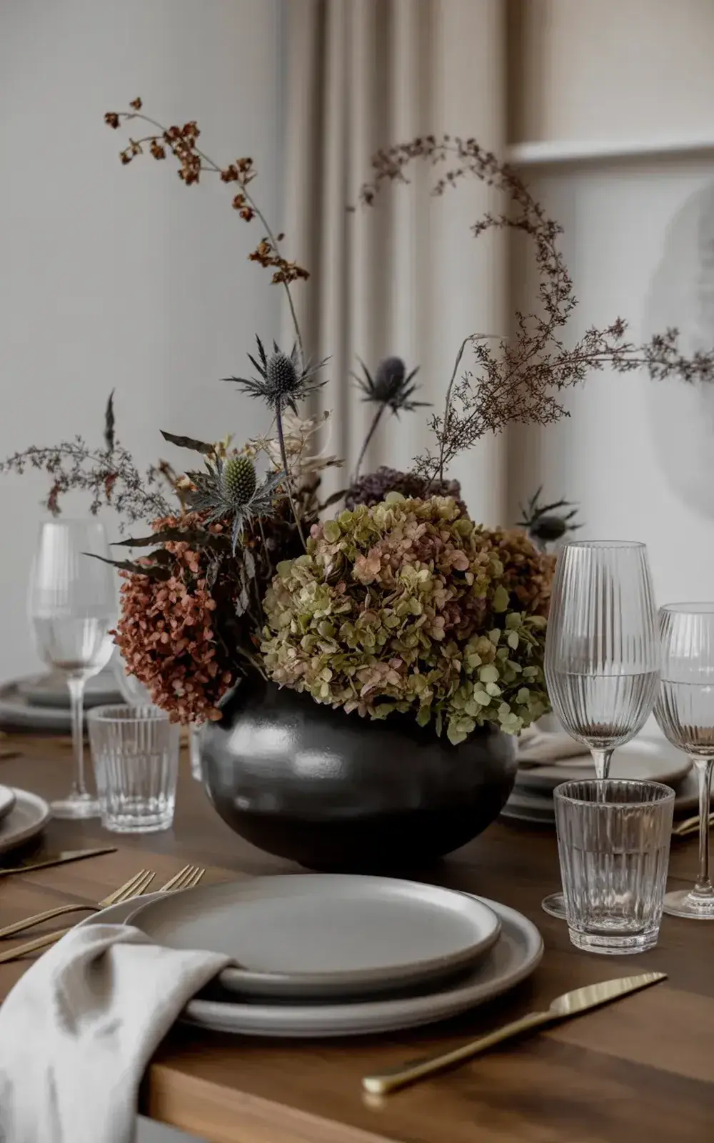 A table set with dark grey floral vase and glassware with white plates and silver cutlery.