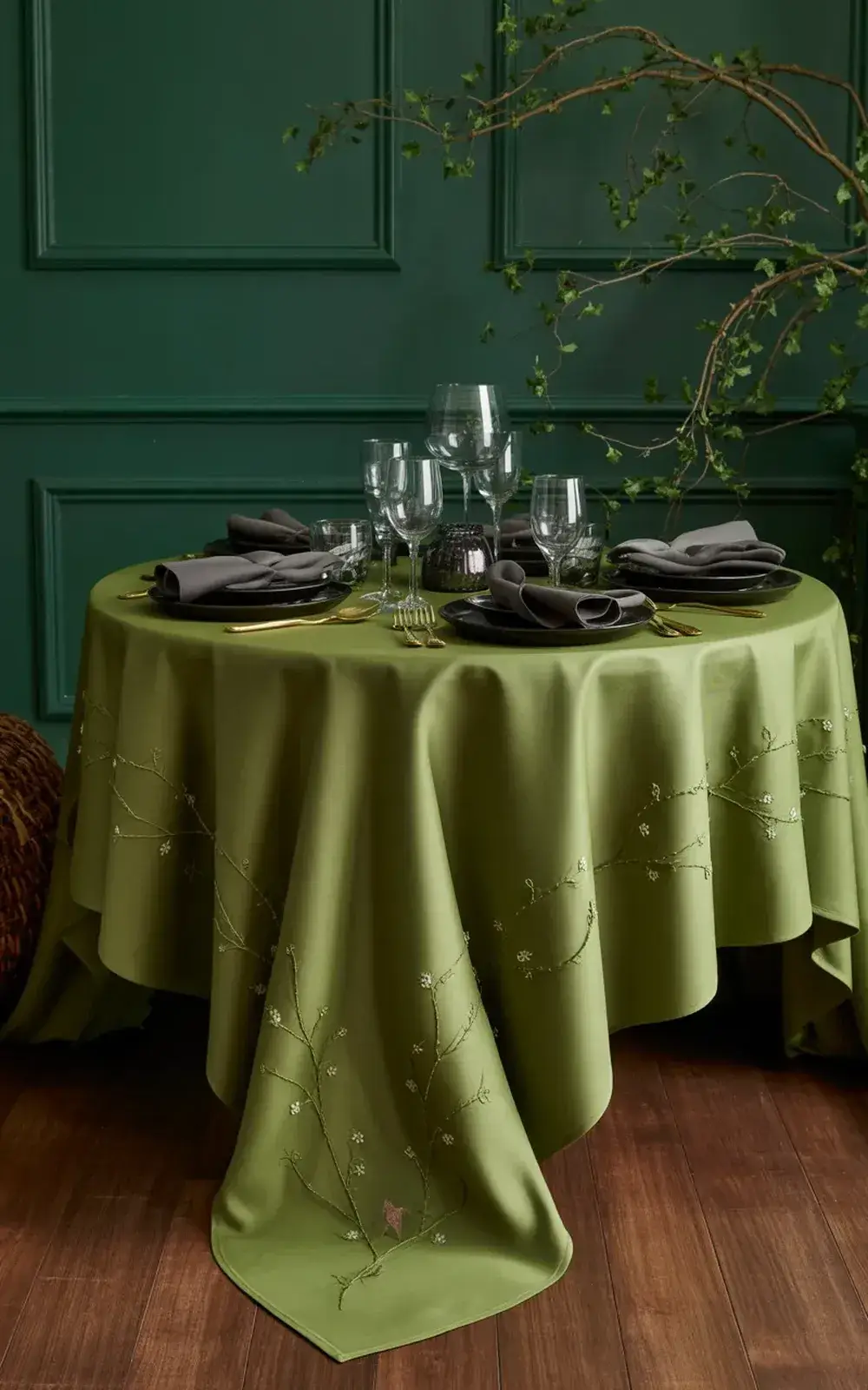 Elegantly set dining table with green tablecloth, glassware, and folded napkins against a dark green wall with greenery.