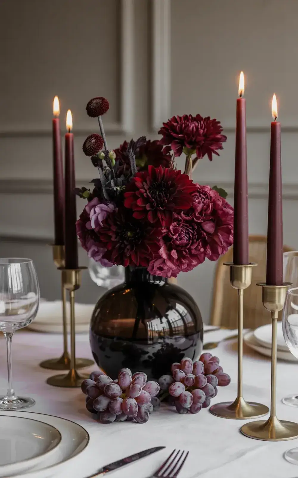 Elegant table setting with a vase of dark red flowers, lit candles, and a cluster of frozen grapes.