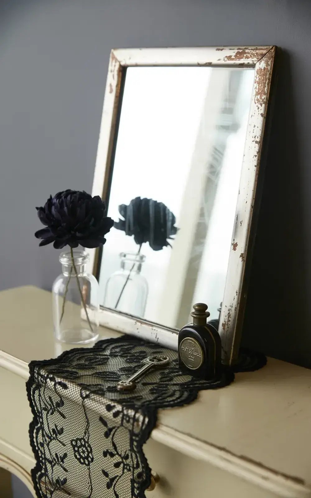 A mirror shows a reflection of black flowers in a glass bottle with lace, bottle, and key surrounding the base.