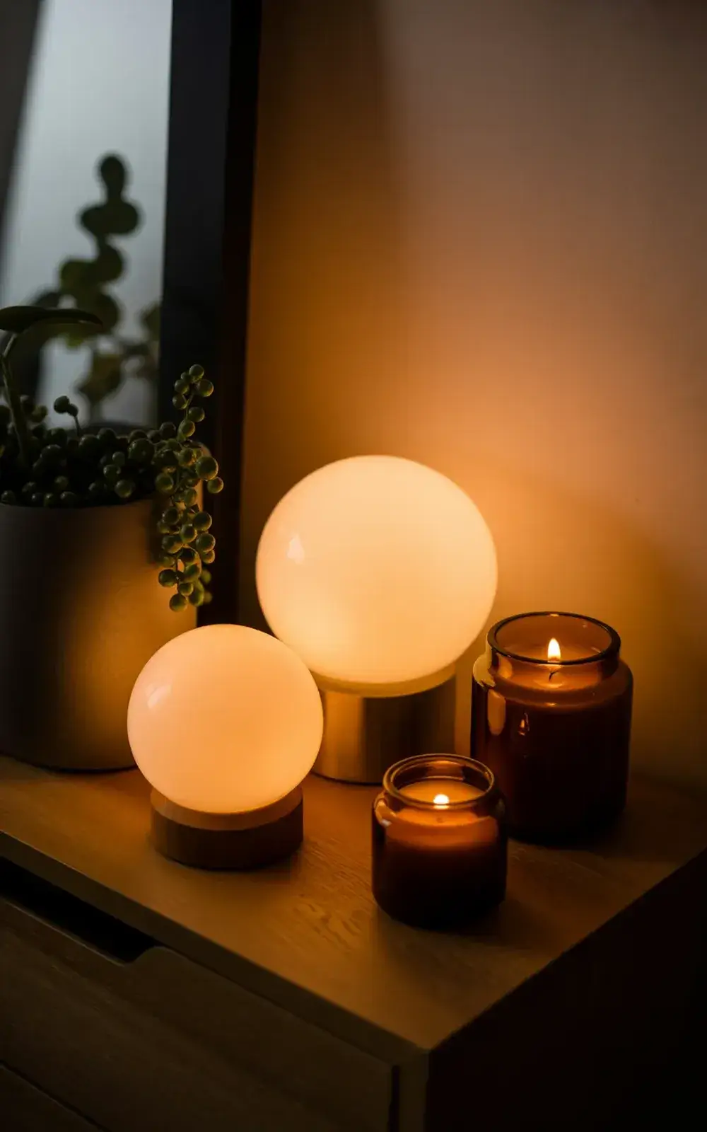 Two lit lamps and two lit candles on a wooden surface near a window with a plant.